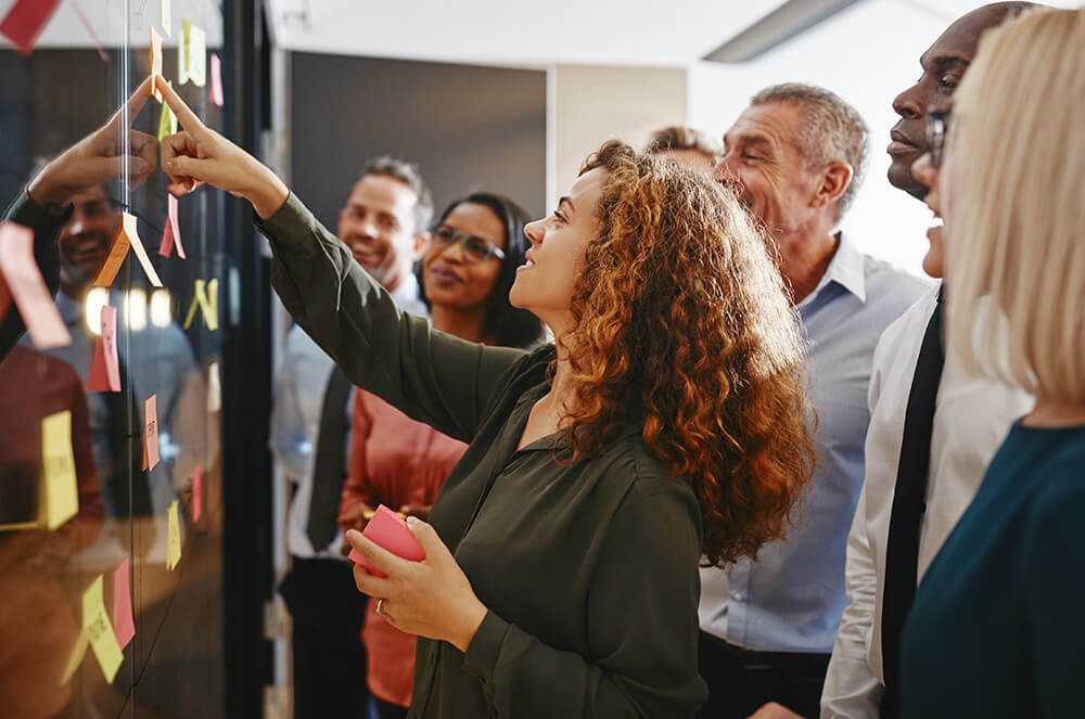 Woman pointing to wall of post-it notes while people watch and smile