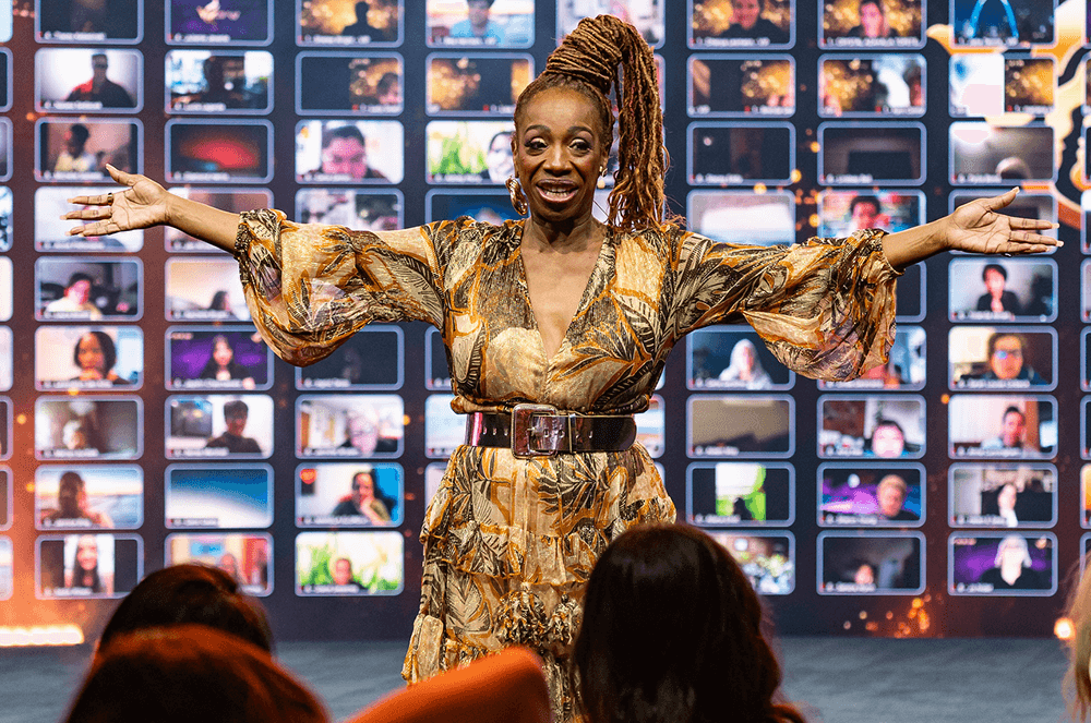 Woman in gold dress speaking onstage with audience members on screen in background