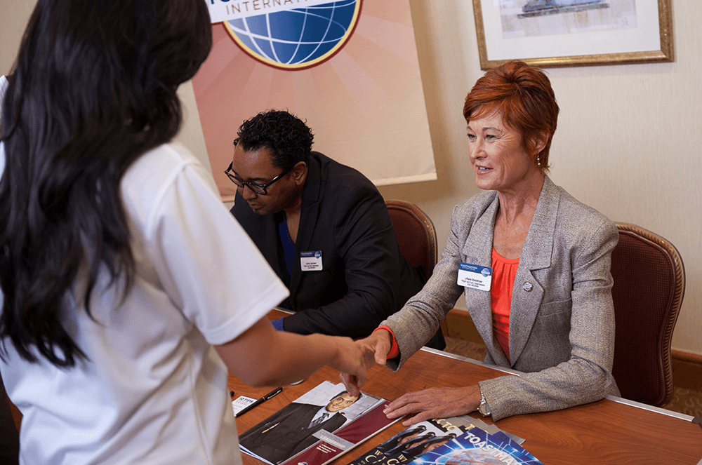 Two women shaking hands at booth with man in background