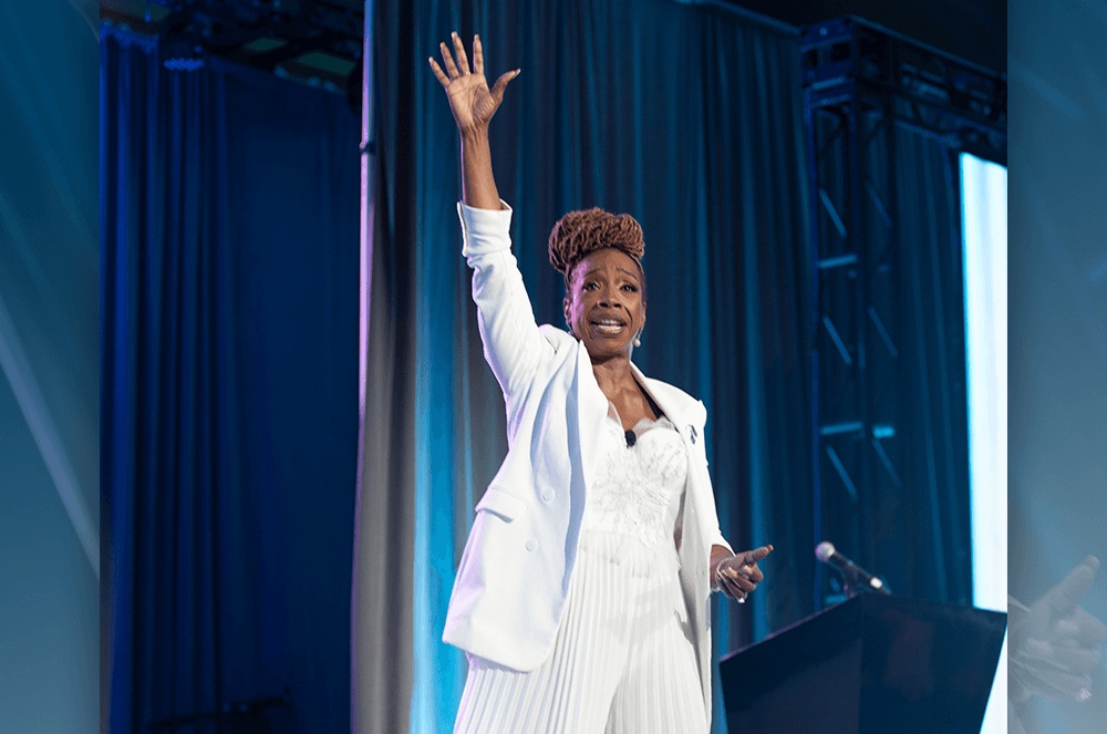 Woman in all white lifting hand up in air while speaking onstage