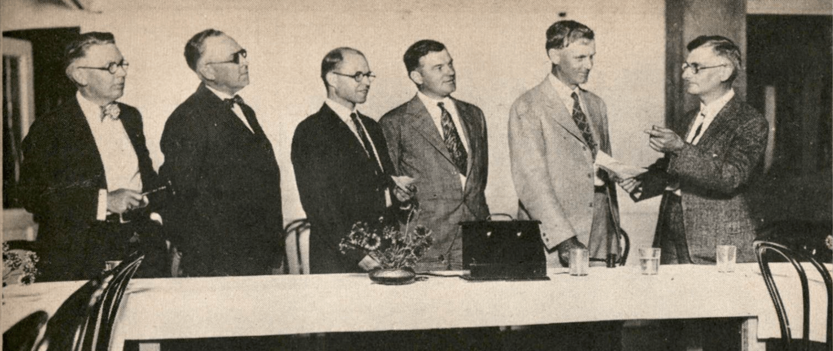 Black and white image of a man leading a meeting with five other men