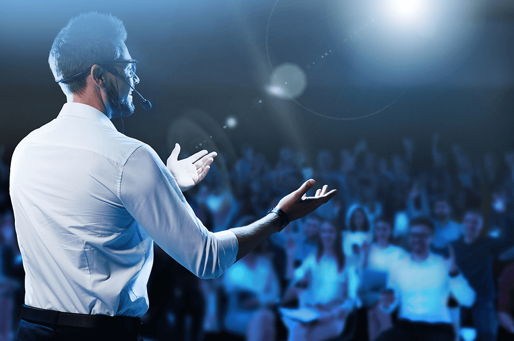 Man onstage with head microphone and hands raised while speaking to audience