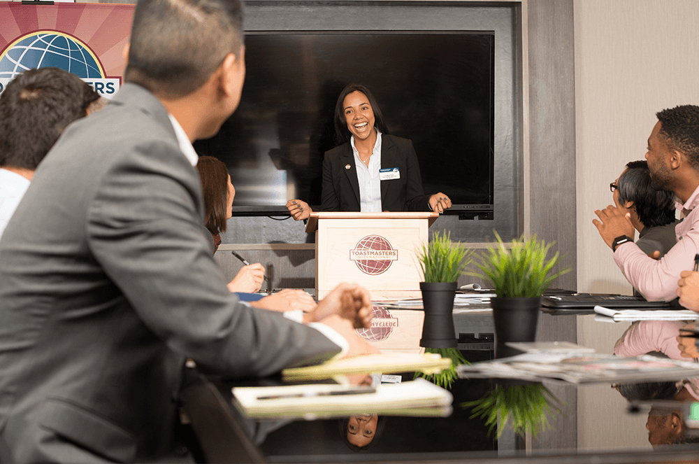 Woman at lectern smiling while members at table laugh