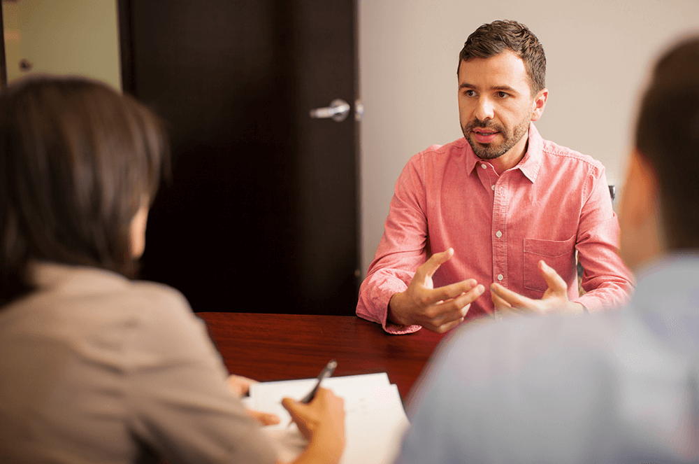 Man in pink shirt being interviewed by man and woman across table