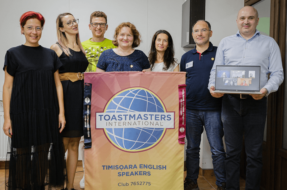 Seven people posing with club banner and laptop with online attendees