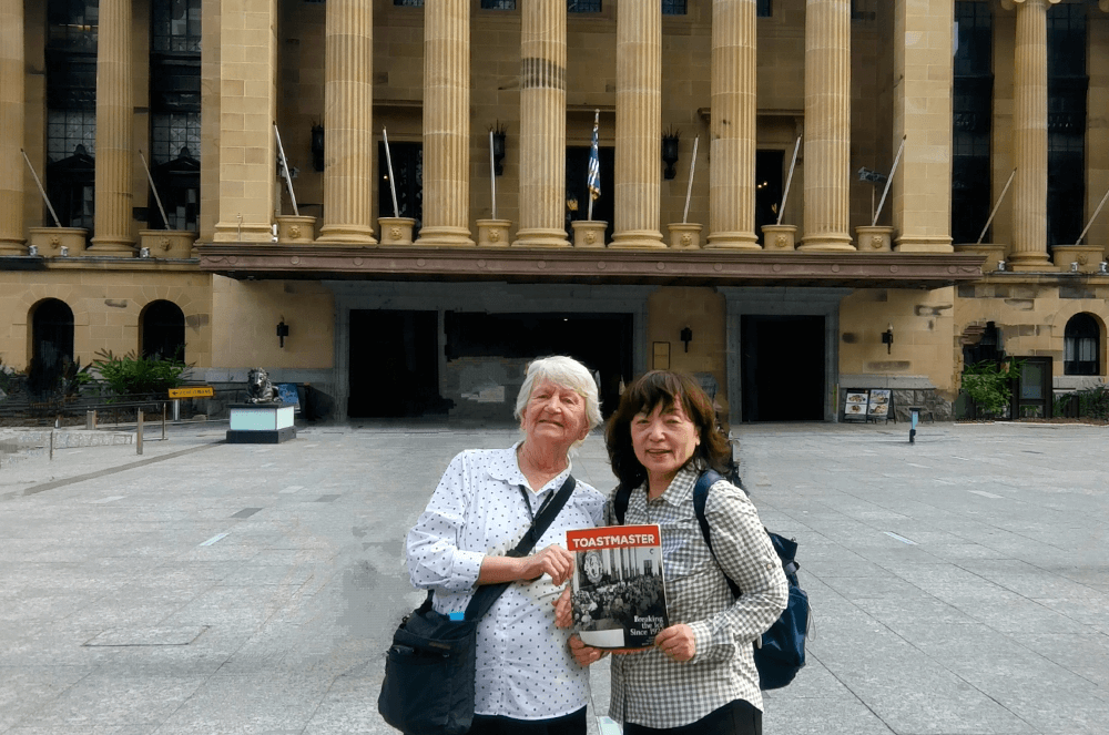 Two women posing with Toastmaster magazine in front of building in Australia