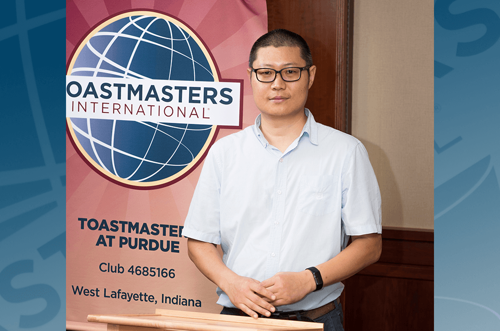 Young man posing at lectern with Toastmasters club banner in background