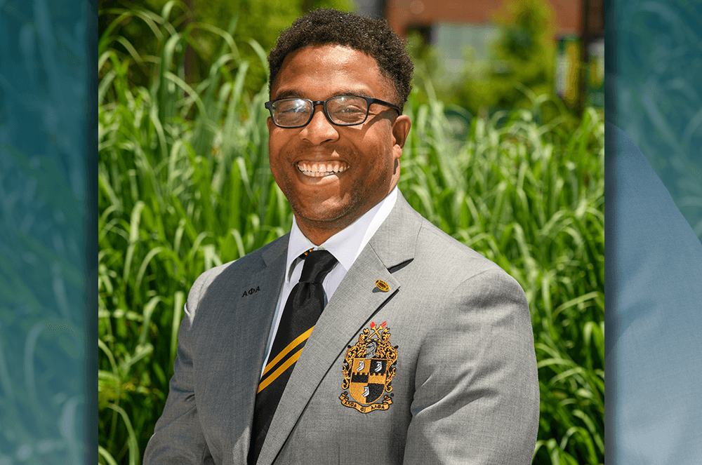 Man in gray suit with collegiate emblem posing outdoors in front of green plants