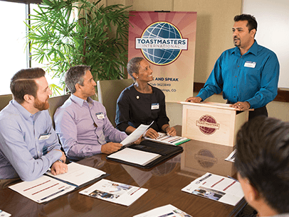 Man speaking at lectern while people listen around table